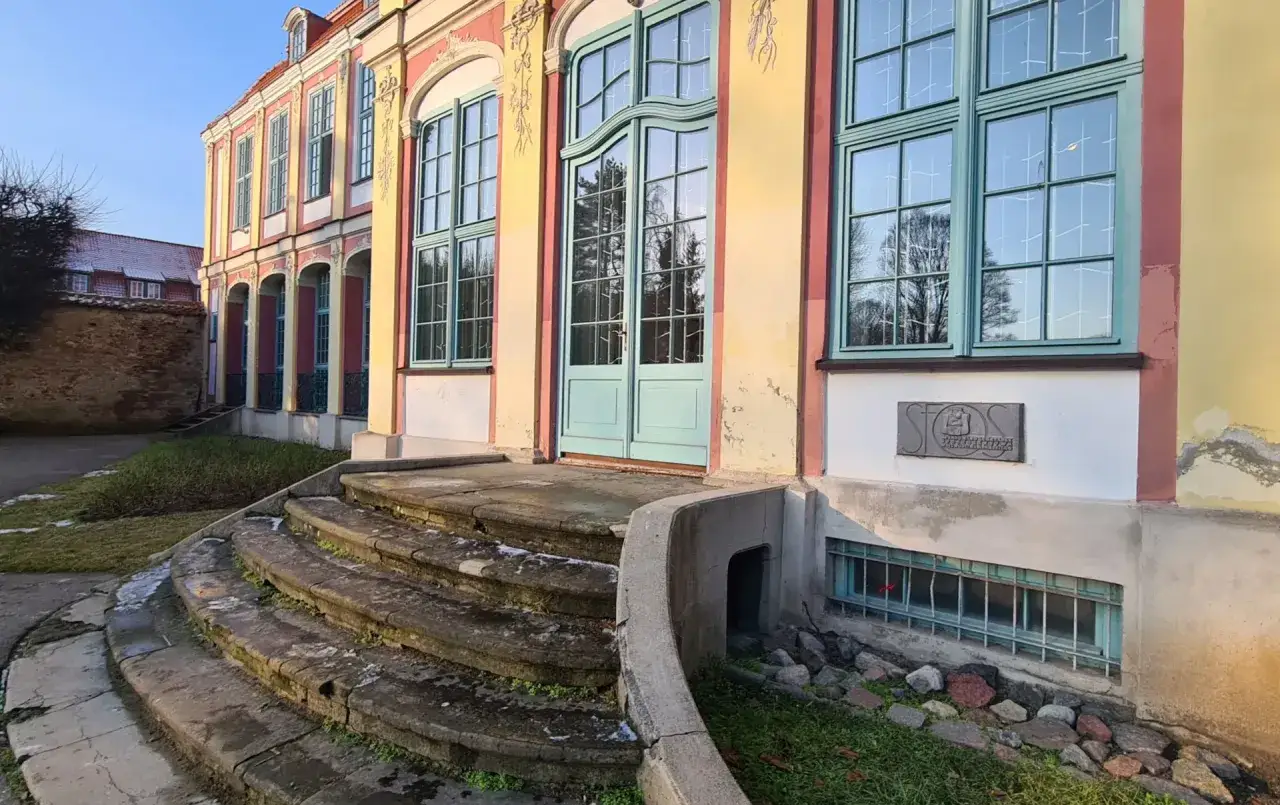 Kamienne schody prowadzą do wejścia pałacu opat&oacute;w w Gdańsku. Budynek ma ż&oacute;łto-r&oacute;żową elewację i zielone okna.