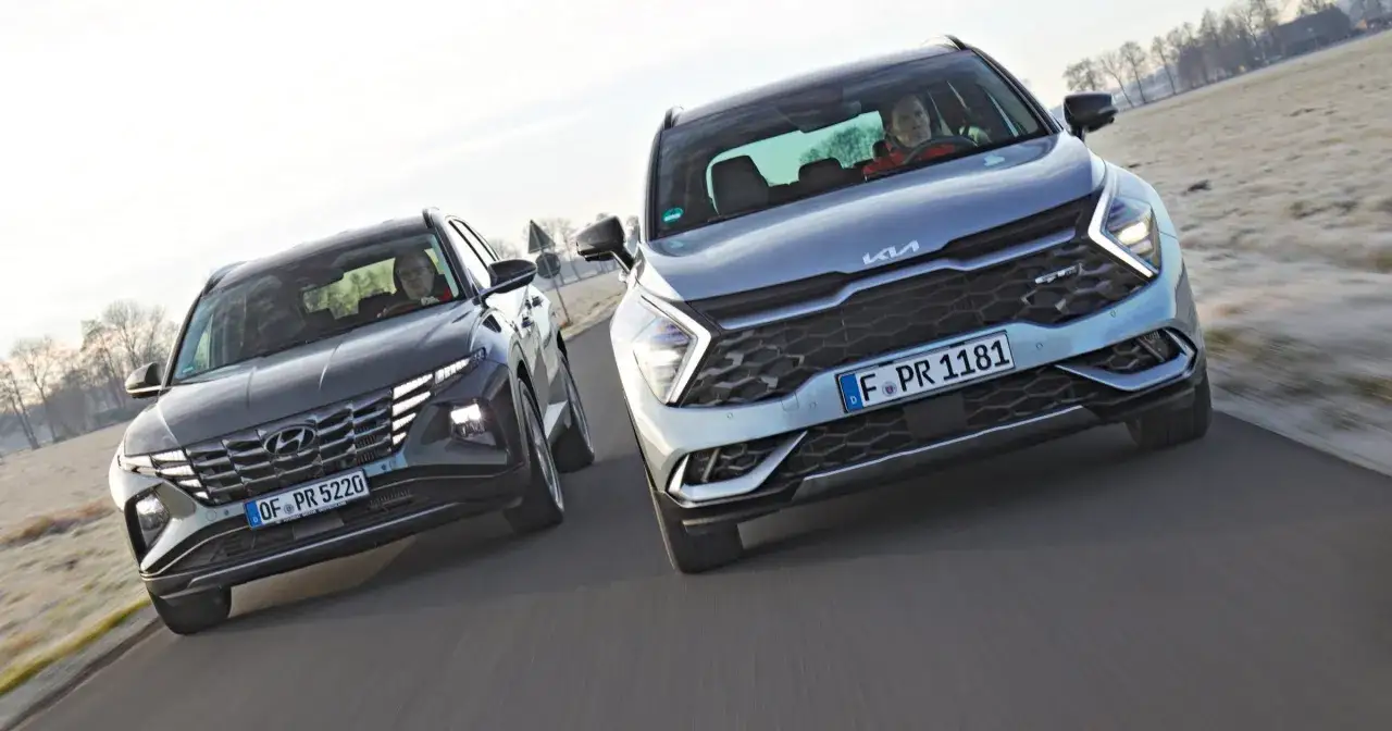 Dwa SUV-y, jeden ciemny Hyundai, drugi jasny **suv kia**, jadą obok siebie po drodze.