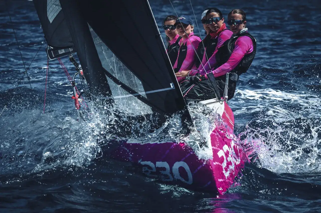 Quattro donne in barca a vela, concentrate e determinate, solcano le onde blu con spruzzi d'acqua scintillanti.