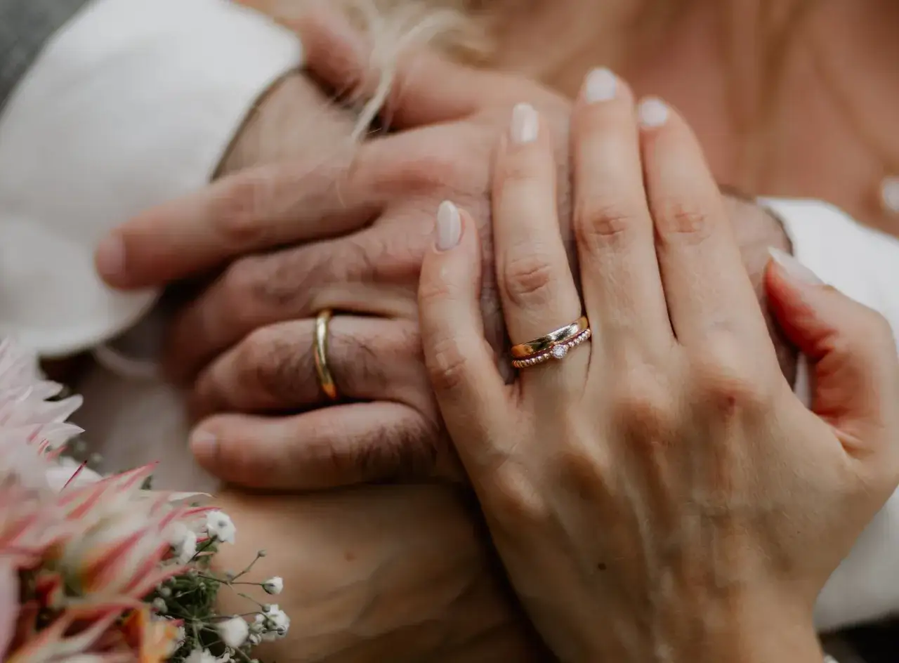 Zwei Hände, die sich halten. Ein Mann trägt einen goldenen ehering, eine Frau trägt zwei zarte Ringe, einer davon mit einem Stein.