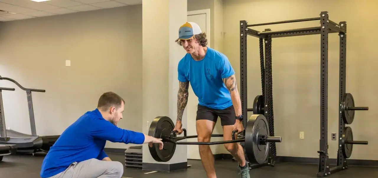 Ein Trainer zeigt einem Mann, wie man mit einer Hantel trainiert. Der Mann mit den Tattoos konzentriert sich auf den Muskelaufbau, während der Trainer ihm hilft.