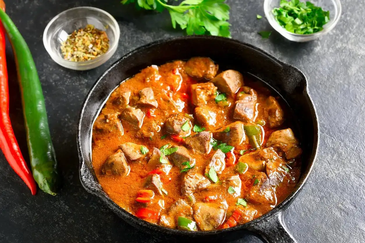 Gulasz z kurczaka w sosie paprykowym, podany w żeliwnej patelni, ozdobiony natką pietruszki. Obok papryczki chili i przyprawy.