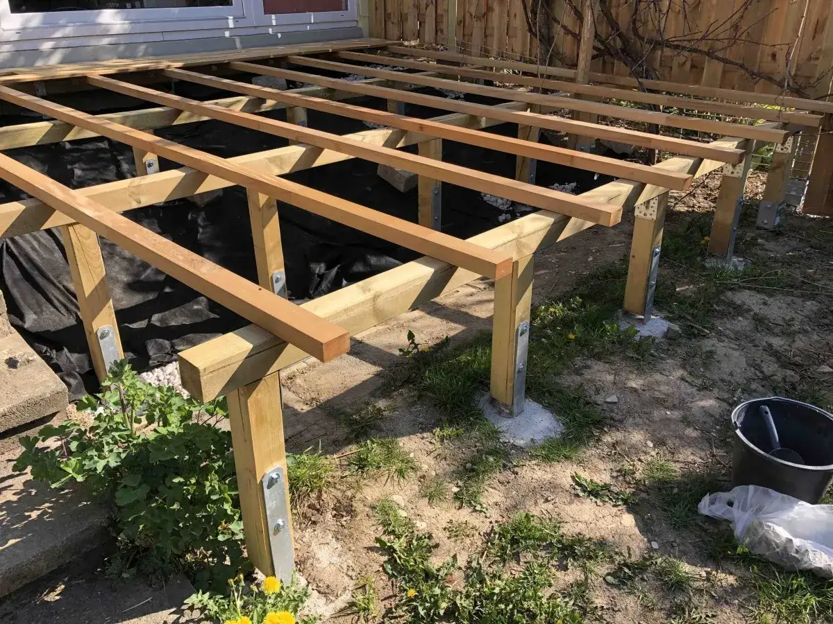 Holzbalken für ein **Podest bauen**. Die Unterkonstruktion steht auf Pfosten, die mit Metallwinkeln befestigt sind. Schwarze Folie schützt den Untergrund.