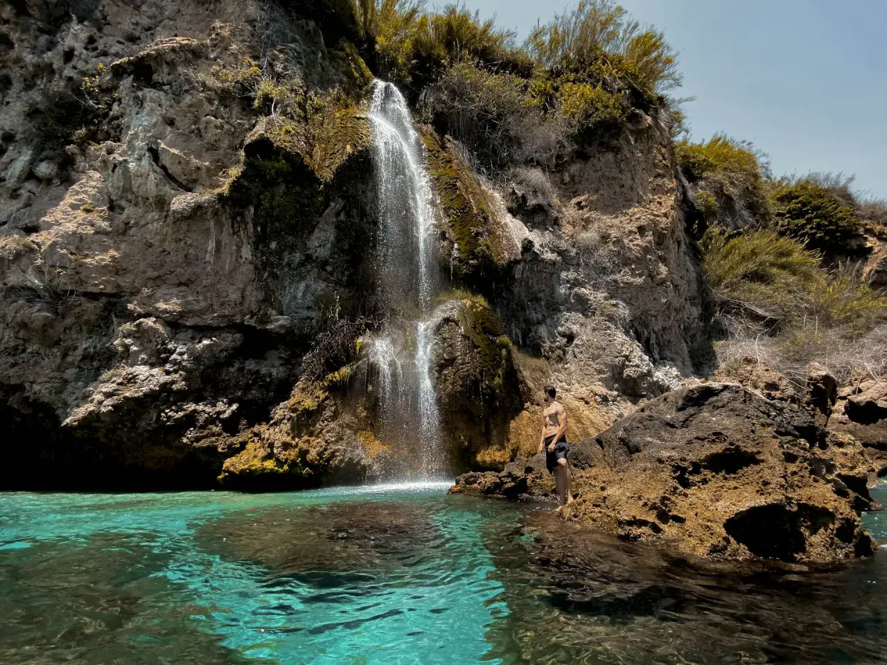 Un joven admira la cascada en la cala barranco de Maro, con aguas turquesas y rocas escarpadas.