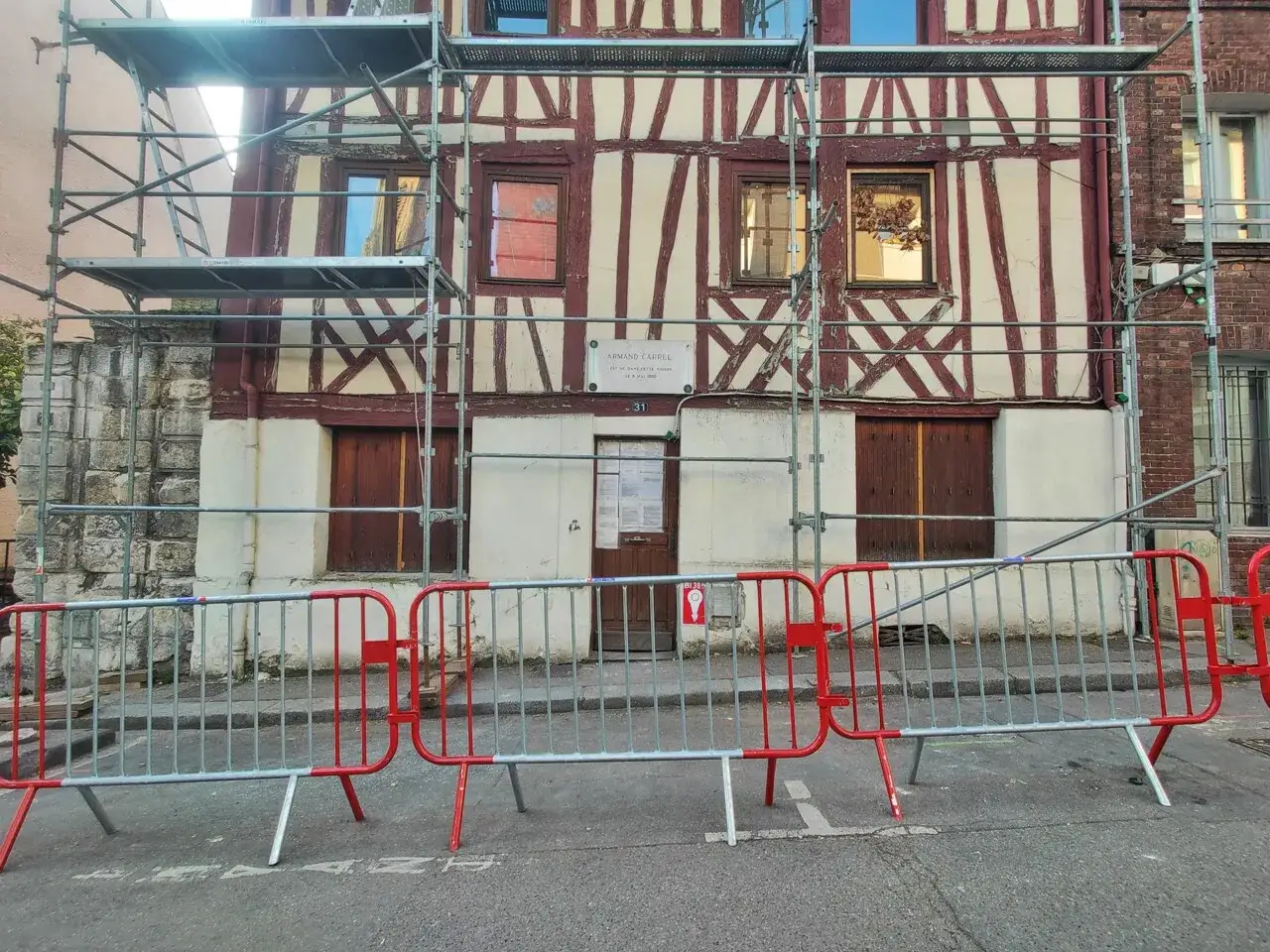 Façade en colombage sous échafaudage, barrières rouges devant. Une plaque indique 