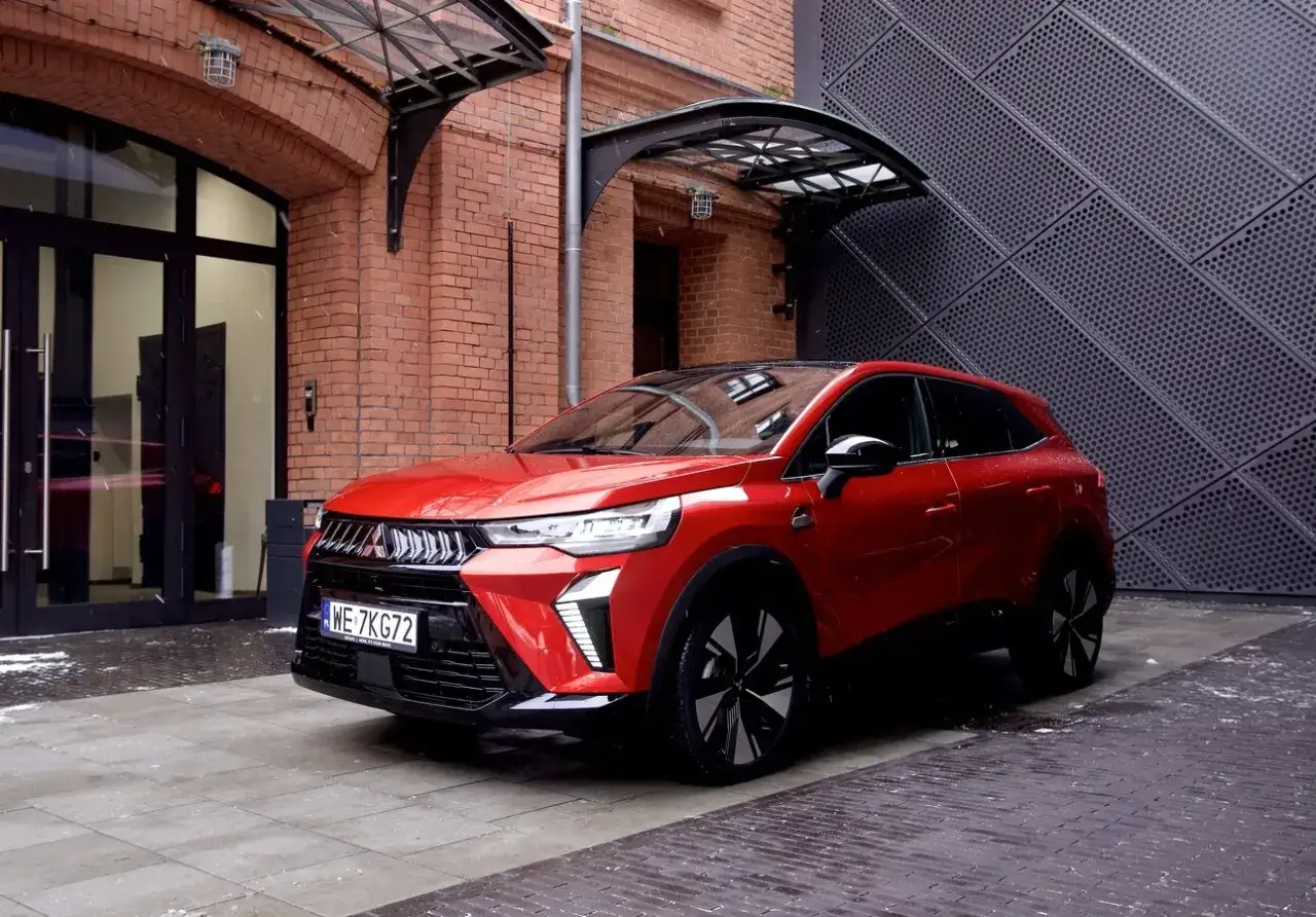 Czerwony SUV, jeden z nowych **mitsubishi modele**, zaparkowany przed budynkiem z cegły i metalową fasadą.