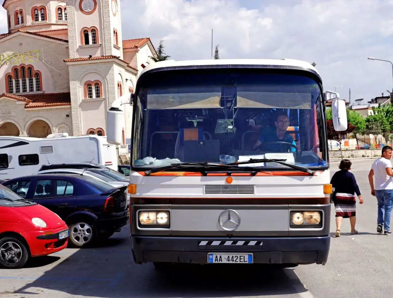 Autobus z kierowcą i tablicą rejestracyjną AL AA 442EL, obok kościół i samochody. To może być wskazówka, jak dojechać do Albanii.