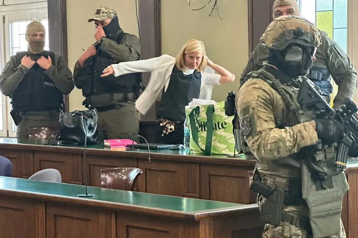 Kobieta w kamizelce kuloodpornej, otoczona przez uzbrojonych funkcjonariuszy, wygląda jak świadek koronny.
