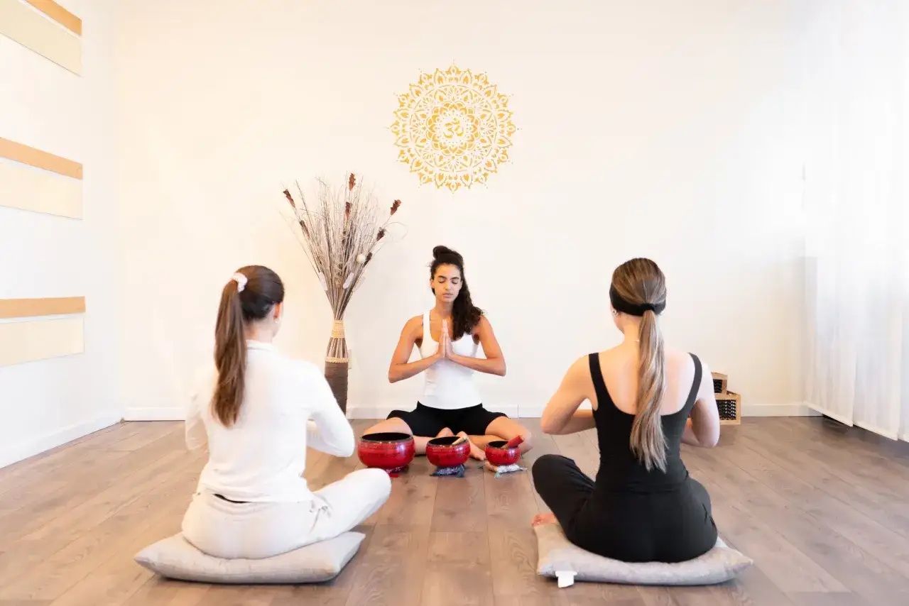 Drei Frauen sitzen im Schneidersitz, üben Pranayama Atmung. Eine goldene Mandalas ziert die Wand.