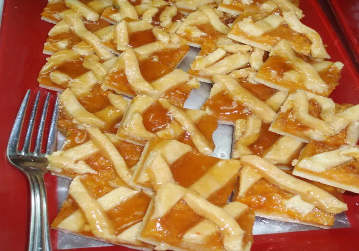 Słodka tarta z brzoskwiniami, pokrojona w kwadraty, z kratką z ciasta. Obok leży widelec.