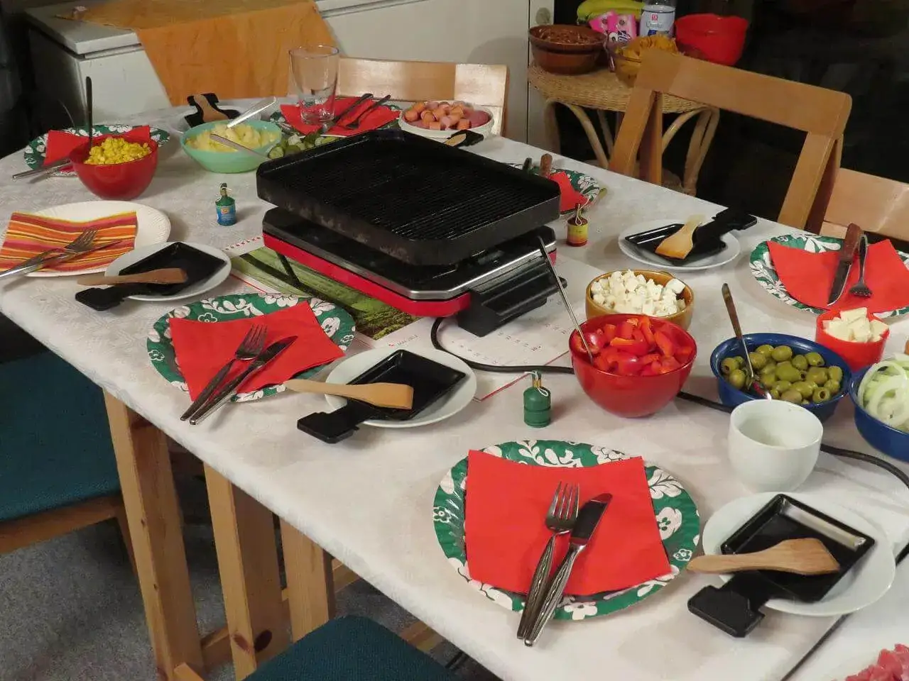 Table dressée pour une raclette sans appareil, avec des bols de fromage, poivrons, olives et maïs.