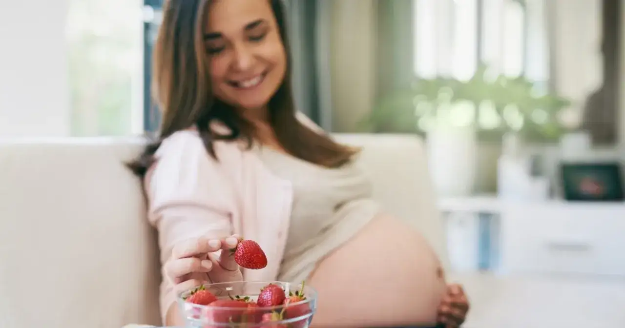Uśmiechnięta ciężarna kobieta trzyma w dłoni truskawkę, obok miseczka pełna owoców. Czas na zdrowe przekąski w ciąży.