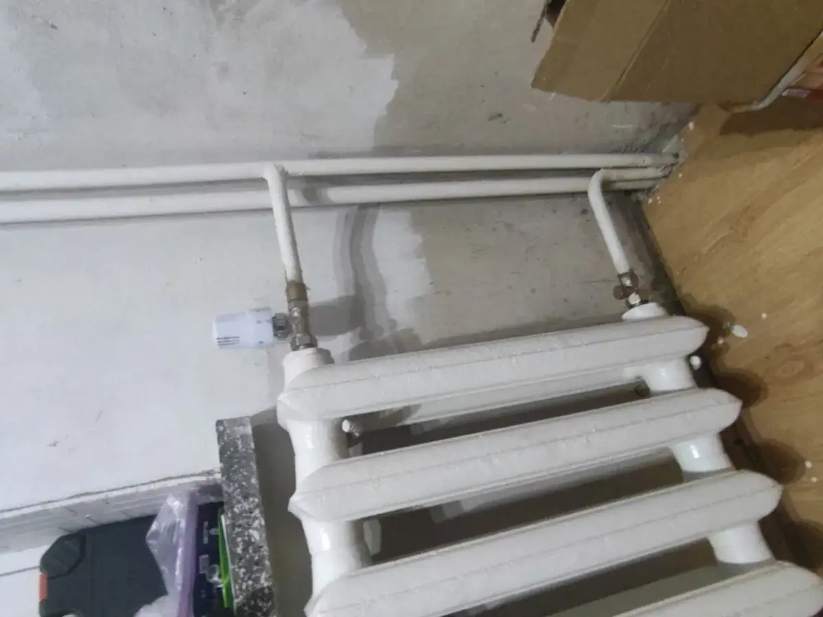 Biały grzejnik łazienkowy podłączony do starej instalacji grzewczej. Widać rury i zawór termostatyczny.