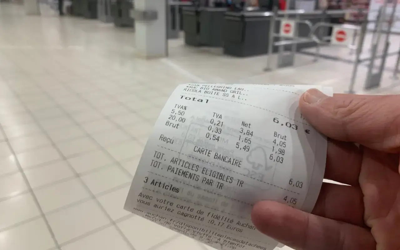 Main tenant un ticket de caisse de supermarché, on voit les articles achetés et le montant total de 6,03 €.