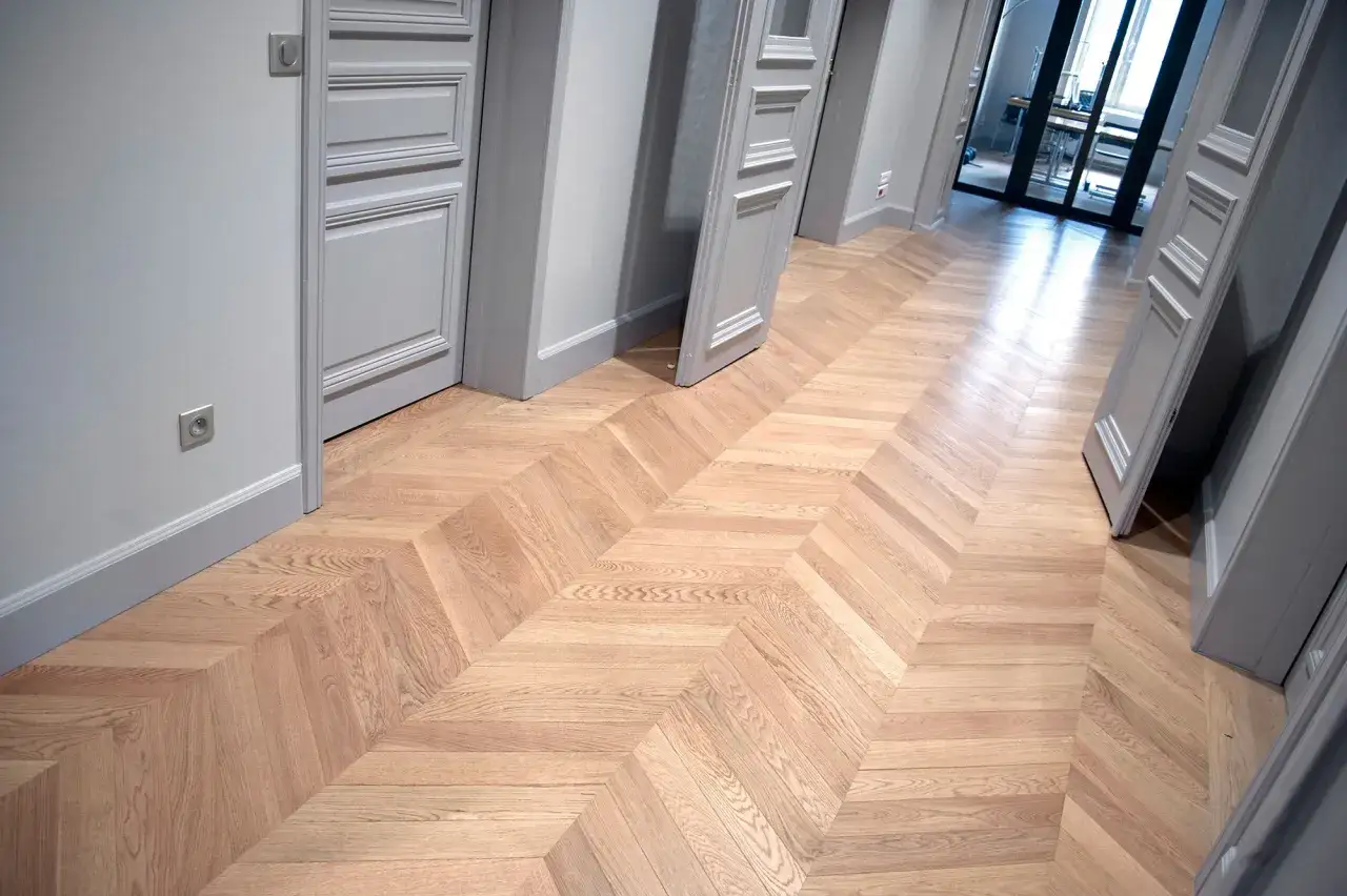 Un couloir &eacute;l&eacute;gant avec un pose parquet point de Hongrie en bois clair. Des portes grises aux moulures fines bordent le passage.
