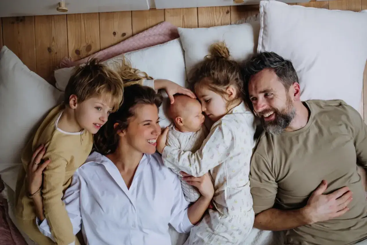 Szczęśliwa rodzina leży razem w łóżku. Mama tuli noworodka, starsze dzieci i tata uśmiechają się. Czas spędzony z rodziną jest bezcenny, jak długi urlop macierzyński.