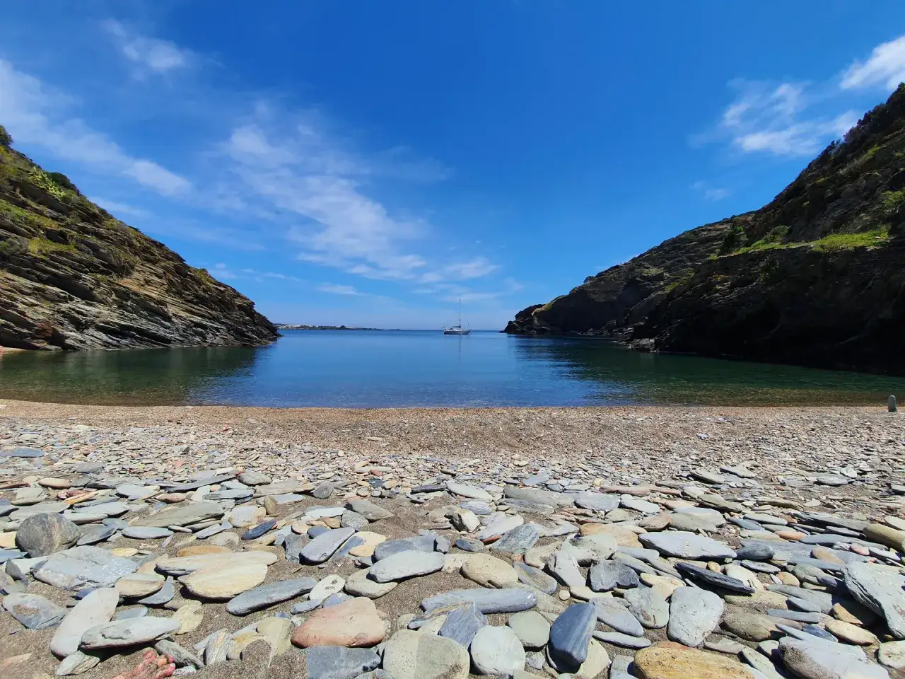 La cala sa sabolla, con su playa de guijarros y aguas cristalinas, invita a la calma. Un velero solitario navega en el horizonte.
