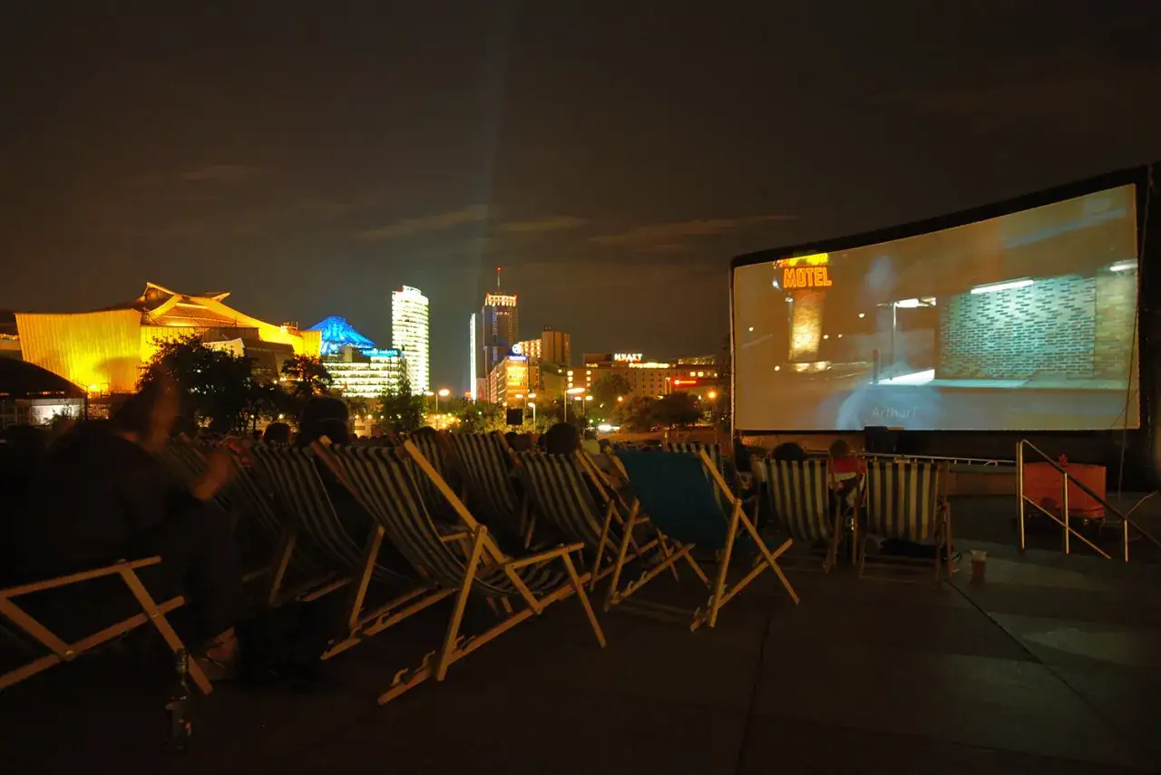 Sommerkino am Potsdamer Platz: Leute schauen Film auf Liegest&uuml;hlen, im Hintergrund beleuchtete Skyline Berlins.