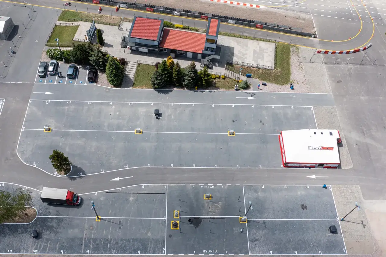 Widok z lotu ptaka na tor kartingowy. Widać budynek z czerwonym dachem, parking z samochodami i ciężarówką, a także fragment toru wyścigowego.