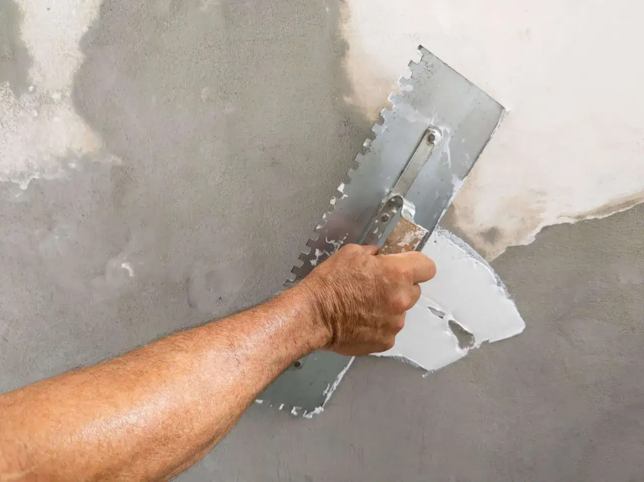 Main d'un artisan appliquant de l'enduit blanc avec une truelle dentel&eacute;e pour enduire un mur ab&icirc;m&eacute;.
