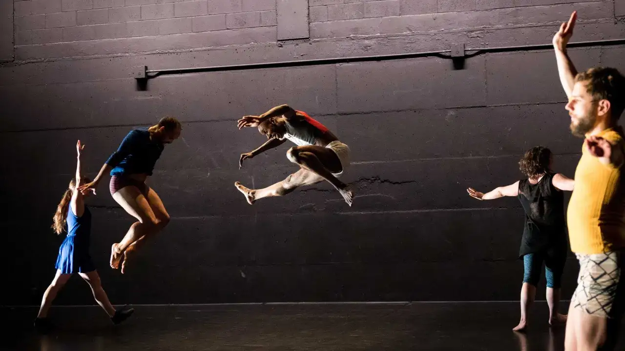 Pięcioro tancerzy wykonuje dynamiczny taniec wsp&oacute;łczesny. Jeden z nich w skoku, pozostali w ruchu.