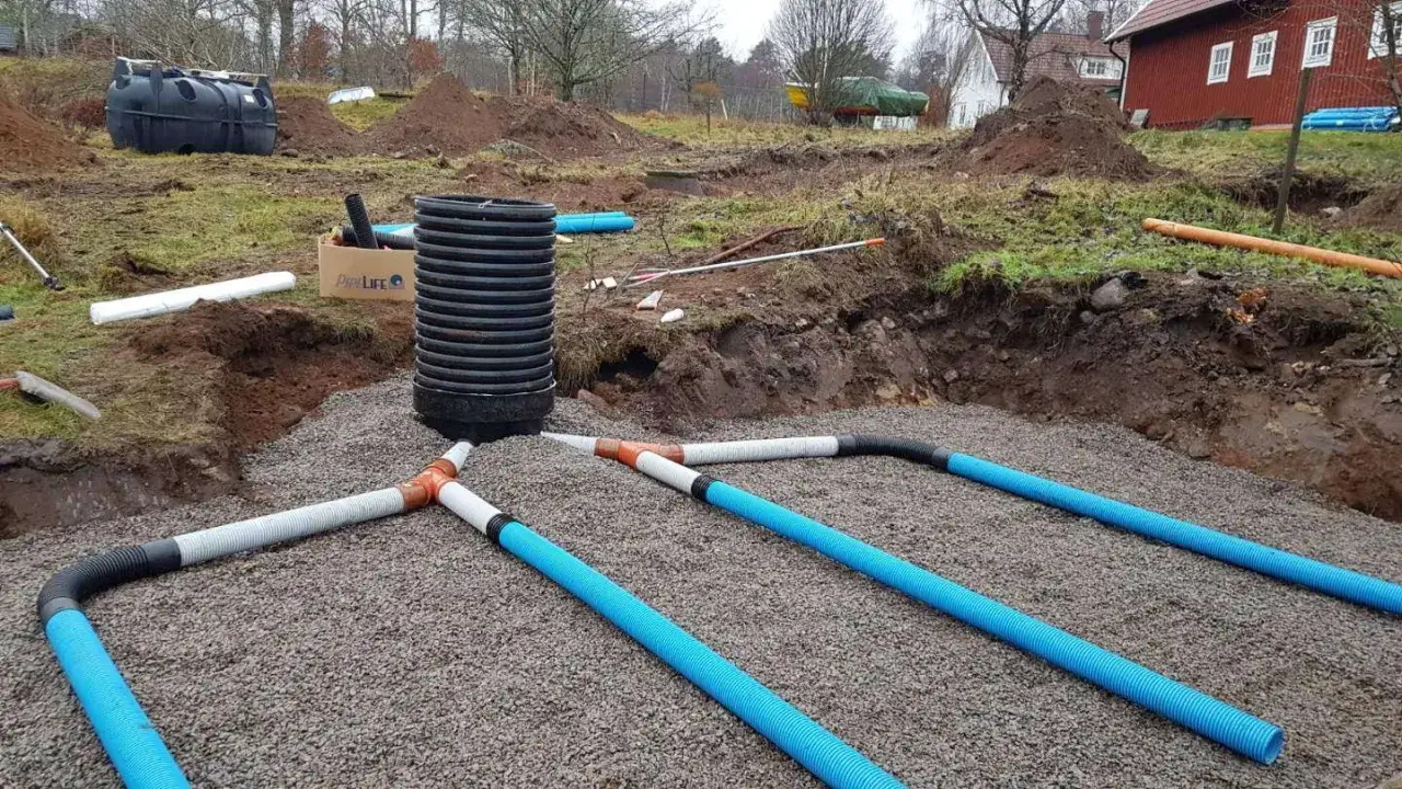 Gråsingel täcker marken där ett enskilt avlopp installeras. Blå och grå rör leder bort vatten från en stor svart tank.