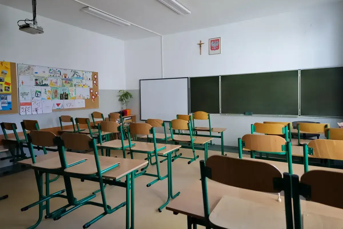 Pusta klasa z rzędami krzeseł i tablicami. W tle rysunki dzieci, symbol Polski i krzyż. To miejsce, gdzie odbywa się doskonalenie zawodowe nauczycieli, a podstawa prawna tego procesu jest kluczowa.
