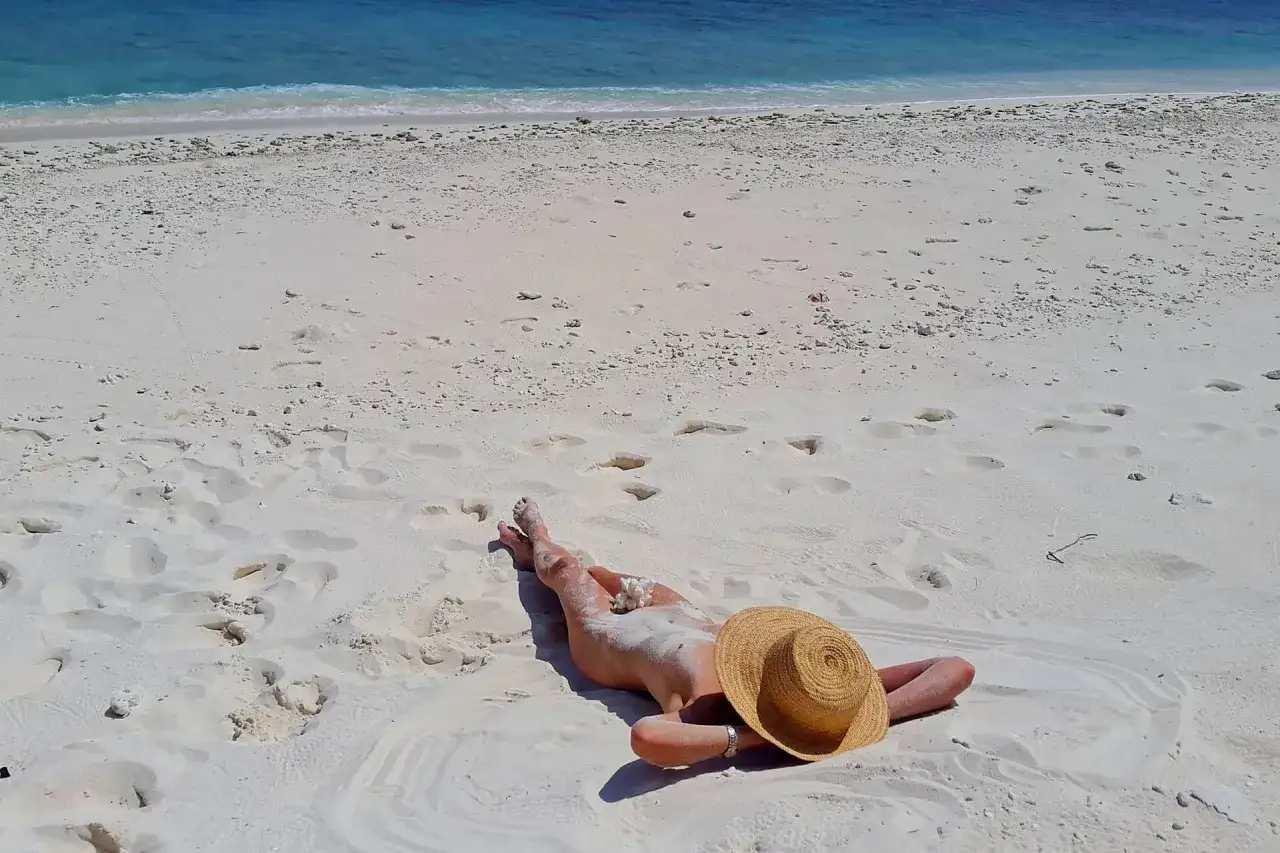 Osoba leży na piaszczystej plaży, opalając się. W tle widać turkusowe morze. To idealne miejsce na plażę nudystów.