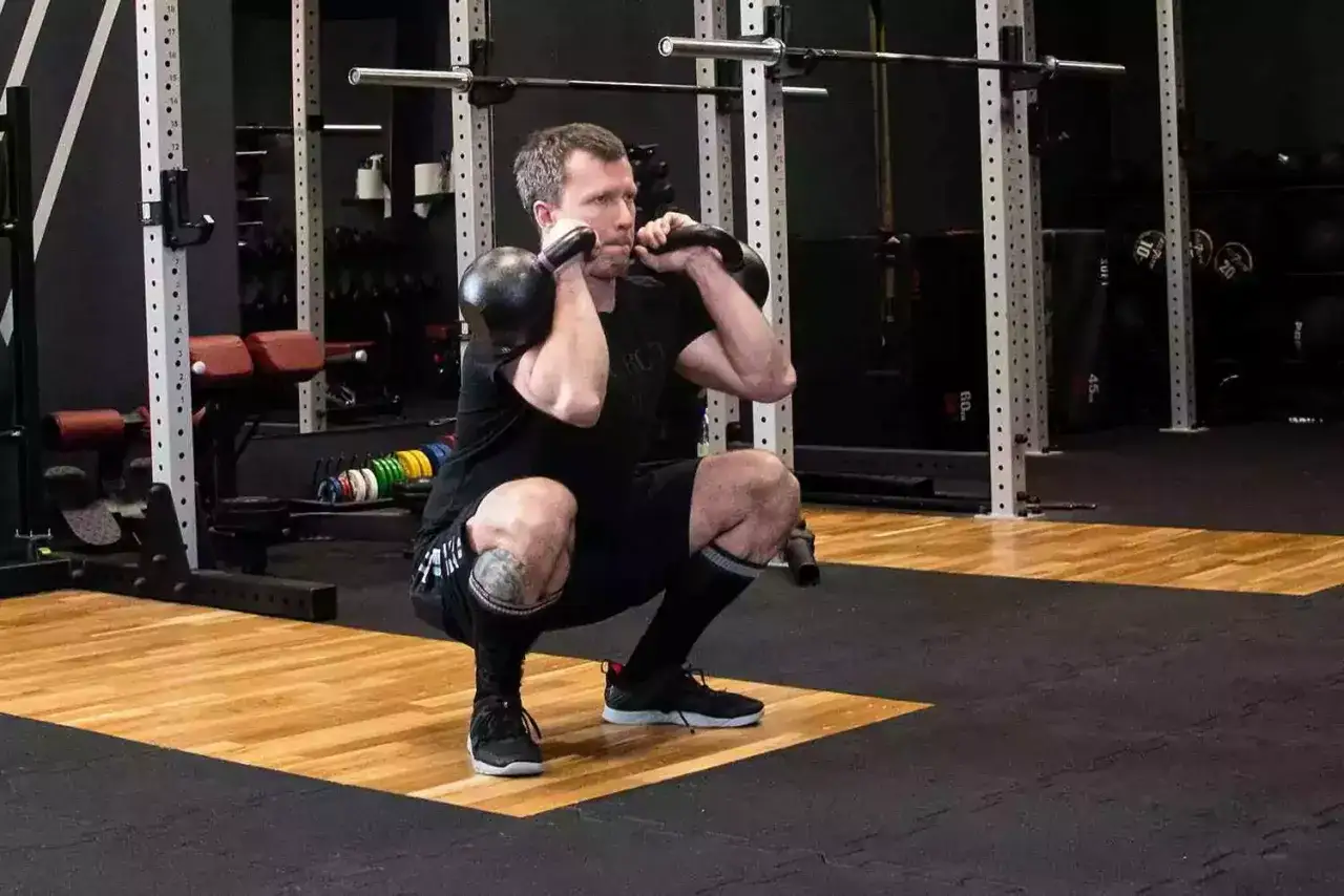 Mężczyzna wykonuje przysiad z kettlebellami. To przykład treningu funkcjonalnego, co to jest? Ćwiczenia poprawiające siłę i koordynację.