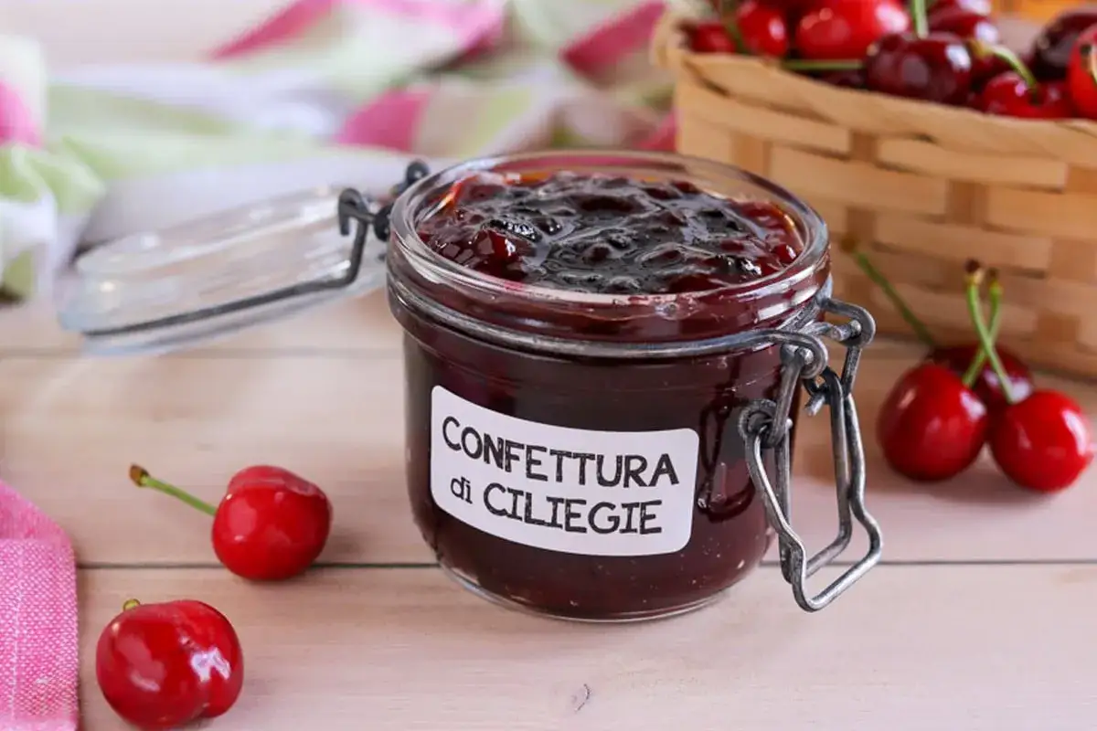 Vaso di confettura di ciliegie fatto in casa, con ciliegie fresche sparse sul tavolo e un cestino sullo sfondo.