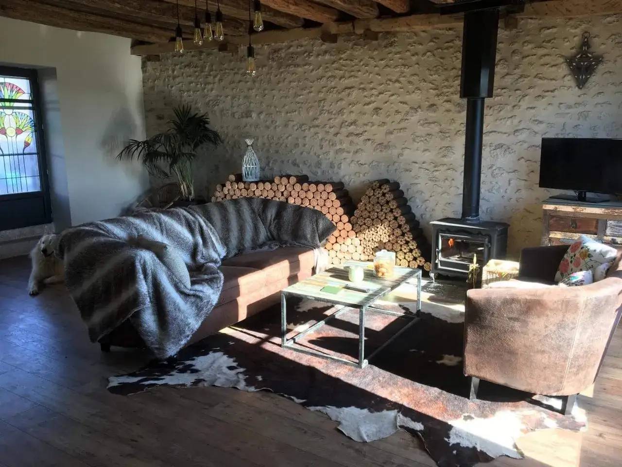 Salon cosy d'une grange en rénovation, avec poêle à bois, canapé confortable et tapis peau de vache.