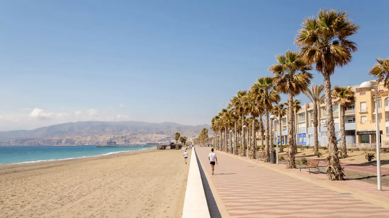 Un paseo mar&iacute;timo en Moj&aacute;car Playa con palmeras, edificios y el mar. Dos personas corren por el carril bici.
