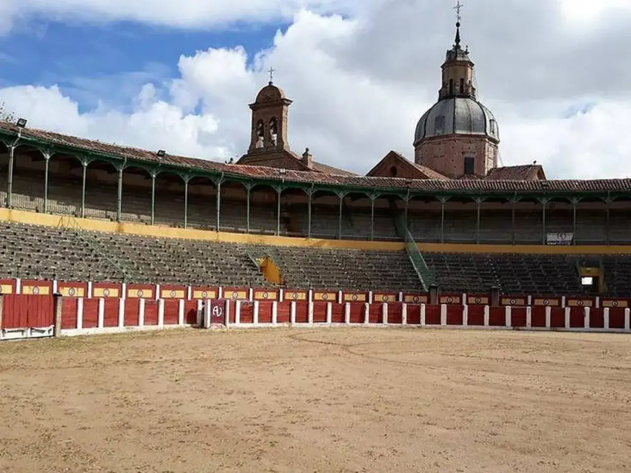 Plaza de toros de Talavera de la Reina, con gradas vac&iacute;as y el campanario de una iglesia al fondo.