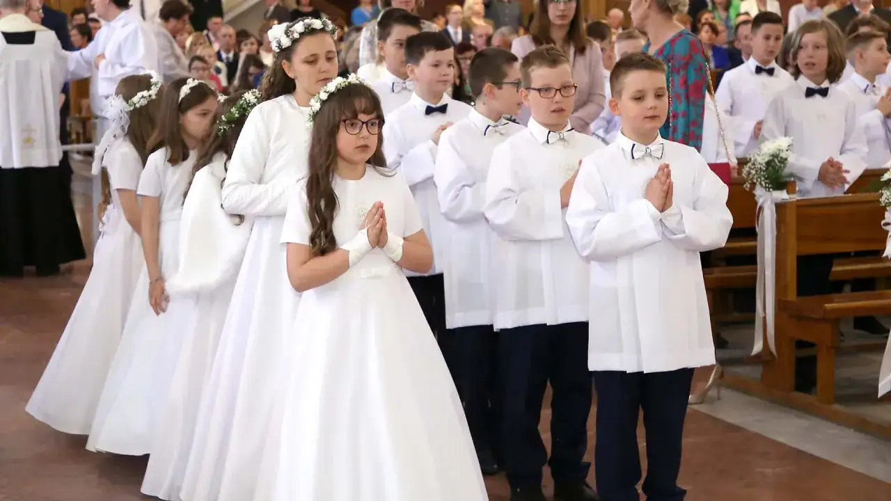 Dzieci w białych strojach, dziewczynki w wiankach, chłopcy w albach, modlą się podczas uroczystości pierwszej komunii świętej.