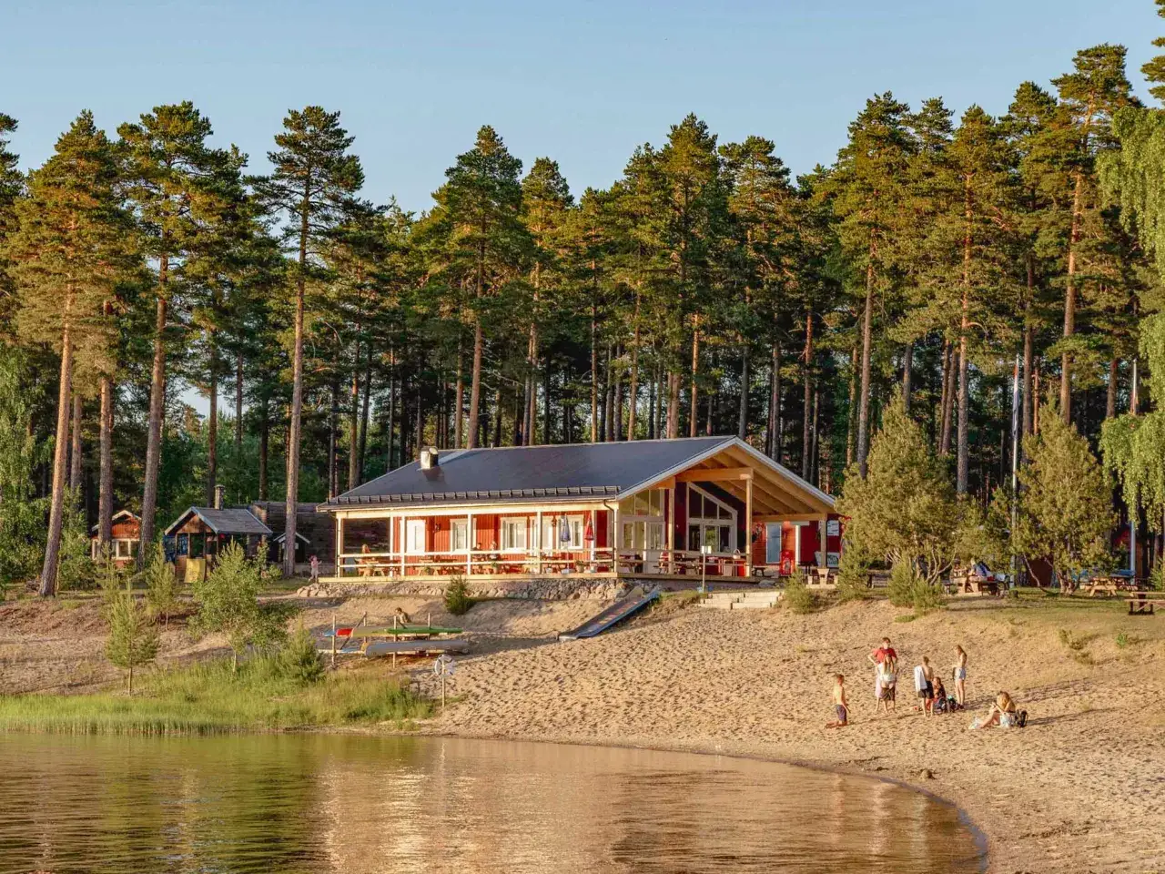 En familj njuter av en solig dag vid sj&ouml;n, redo att campa med barn. En r&ouml;d byggnad med veranda och en sandstrand kantad av tallar.