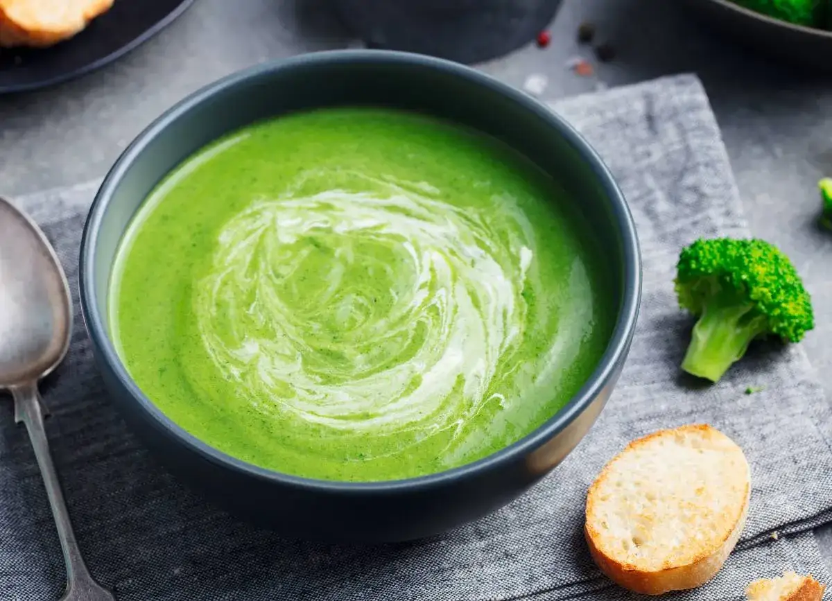Un bol de velouté de brocolis, onctueux et crémeux, avec un tourbillon de crème. Une tête de brocoli et une tranche de pain grillé sont à côté.