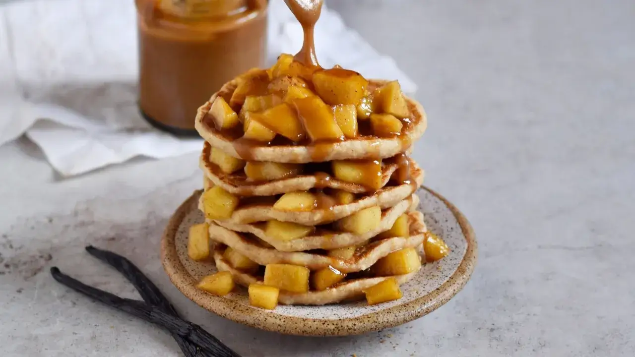 Ein Stapel fluffiger Pfannkuchen mit Hafermilch, belegt mit karamellisierten Äpfeln und Sauce. Daneben liegen Vanilleschoten.