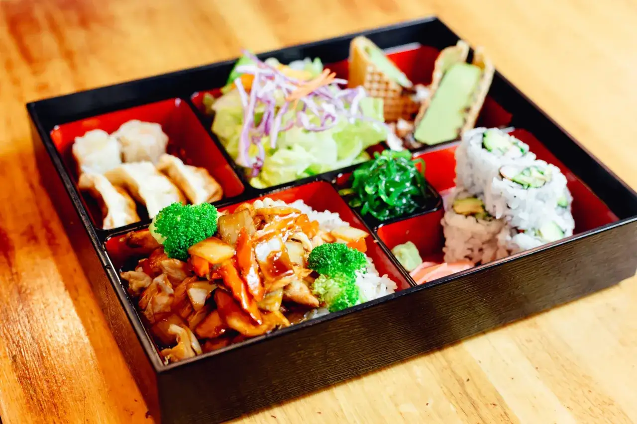 A delicious chicken bento box with sushi, gyoza, salad, and seaweed salad.