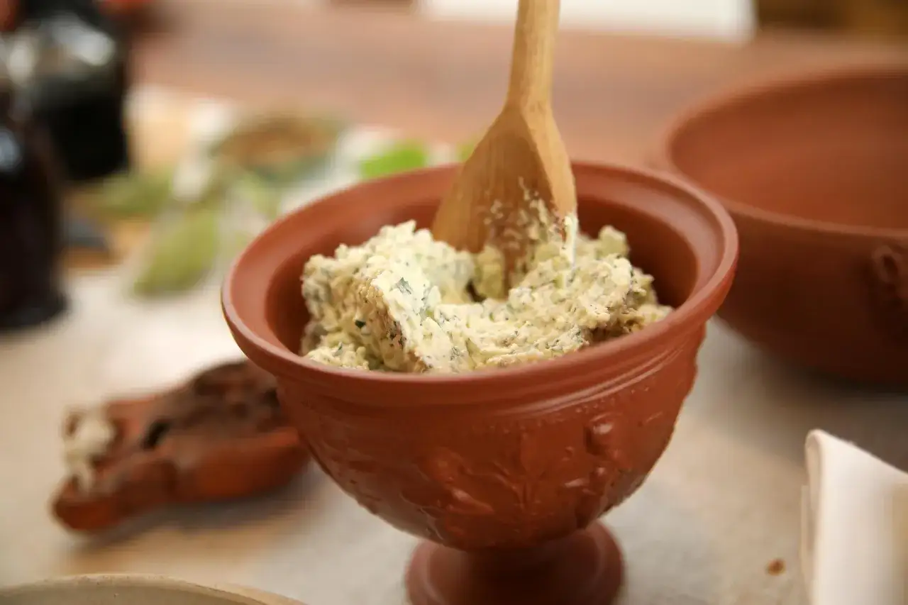 Ein rustikales Tongefäß mit cremigem Moretum, einem Kräuter-Käse-Dip, und einem Holzspatel. Perfekt für ein antikes Moretum Rezept.