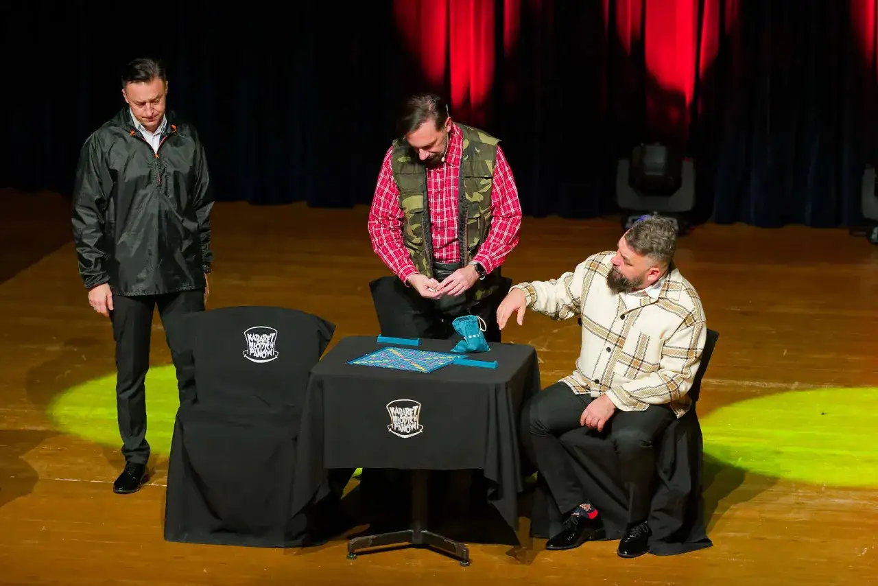 Trzech panów na scenie, jeden stoi, dwóch siedzi. Na stole gra, a na krzesłach koszulki z logo Kabaret Młodych Panów.