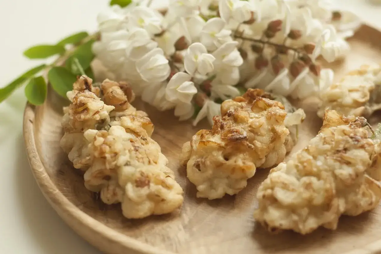Fiori di acacia fritti dorati su un piatto di legno, con grappoli di fiori freschi sullo sfondo.