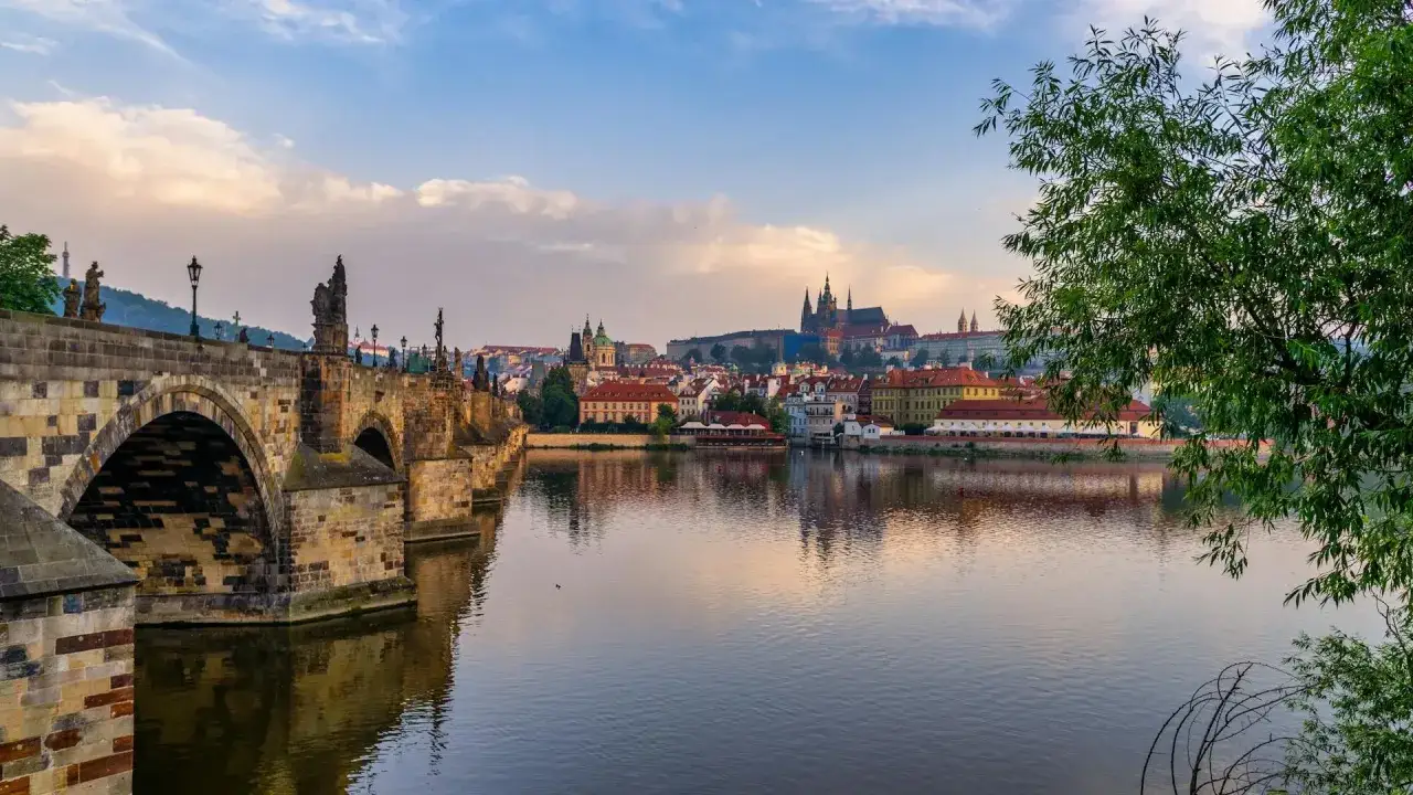 Kamienny most w Czechach, z rzeźbami i latarniami, odbija się w spokojnej rzece. W tle widać panoramę miasta z zamkiem.