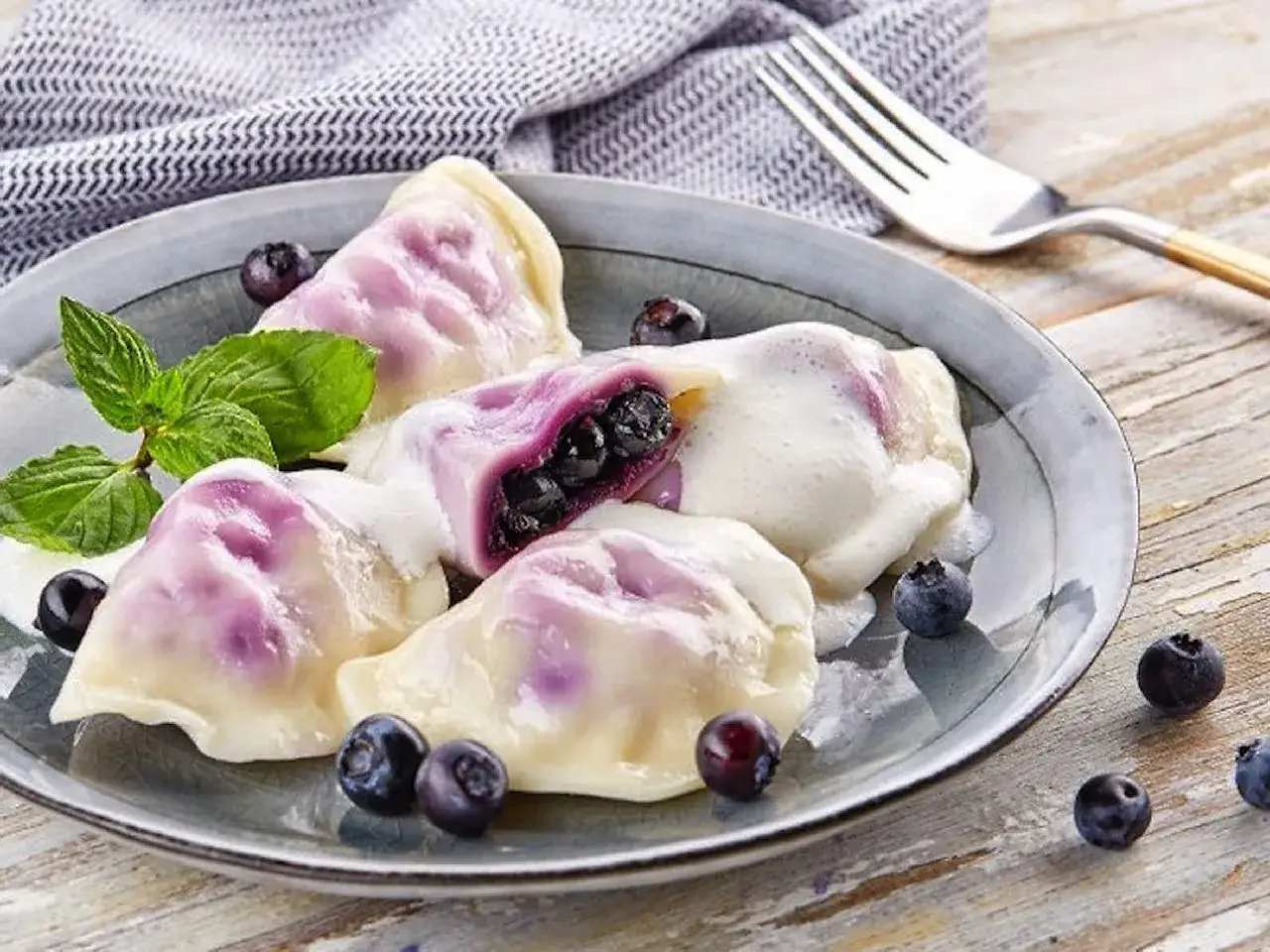 Pyszne pierogi z jagodami, polane śmietaną, ozdobione miętą i świeżymi owocami. Idealne na deser.