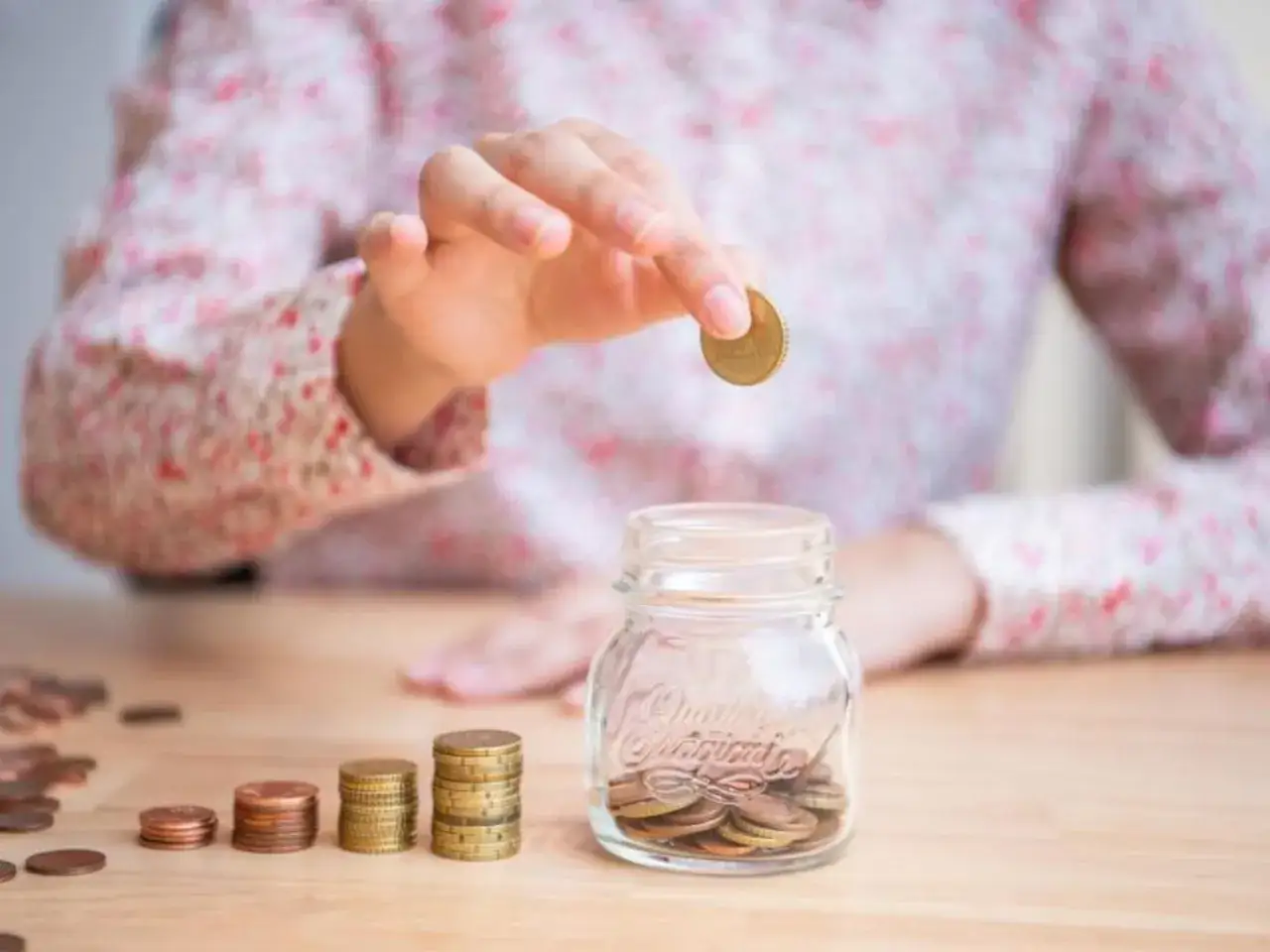Eine Hand wirft eine Münze in ein Sparschwein. Stapel von Münzen liegen daneben. So spart man am besten Geld für die Zukunft.