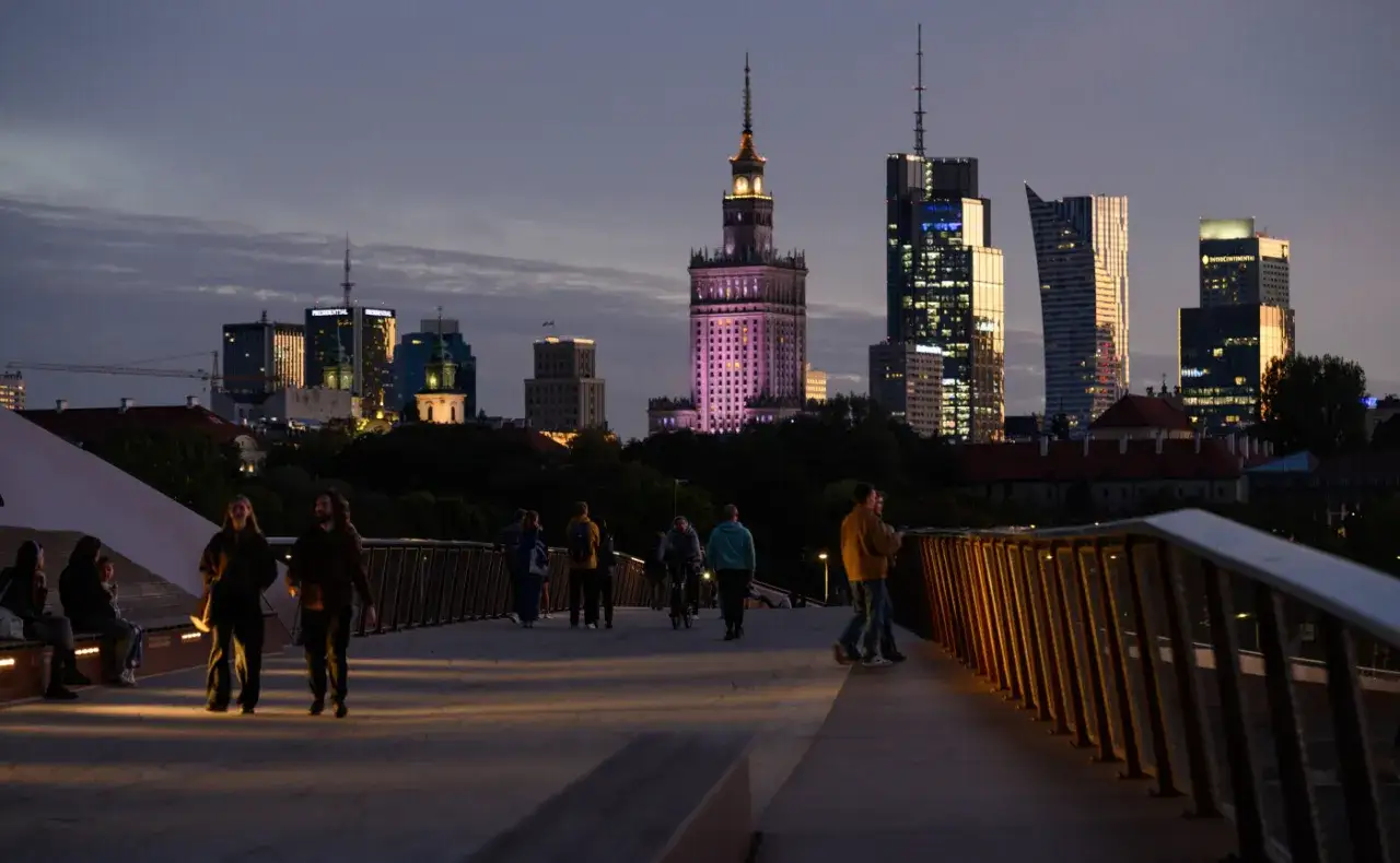 Abendliche Skyline von Warschau, Polen. Menschen spazieren auf einer Br&uuml;cke, w&auml;hrend im Hintergrund beleuchtete Wolkenkratzer und der Kulturpalast erstrahlen. Ein Blick auf die Zukunft mit ETF Polen.