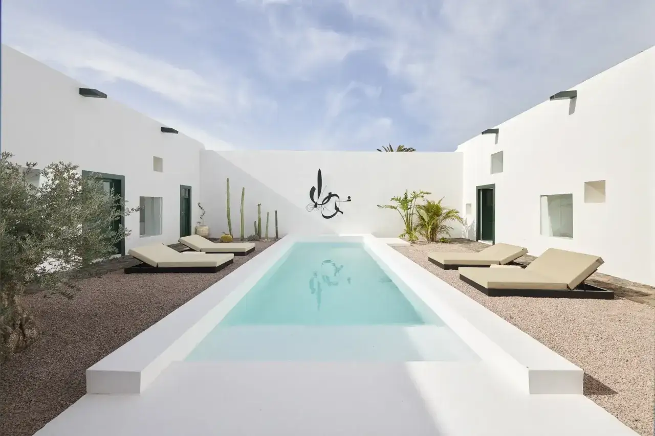 Un oasis moderno con una piscina blanca, tumbonas y cactus bajo un cielo azul.