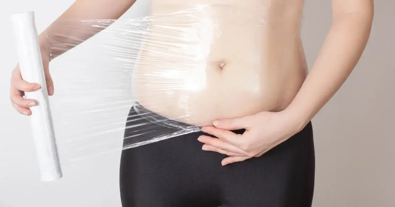 Osoba owija brzuch folią spożywczą, przygotowując się do zabiegu body wrapping. Efekty mogą być widoczne po kilku sesjach.
