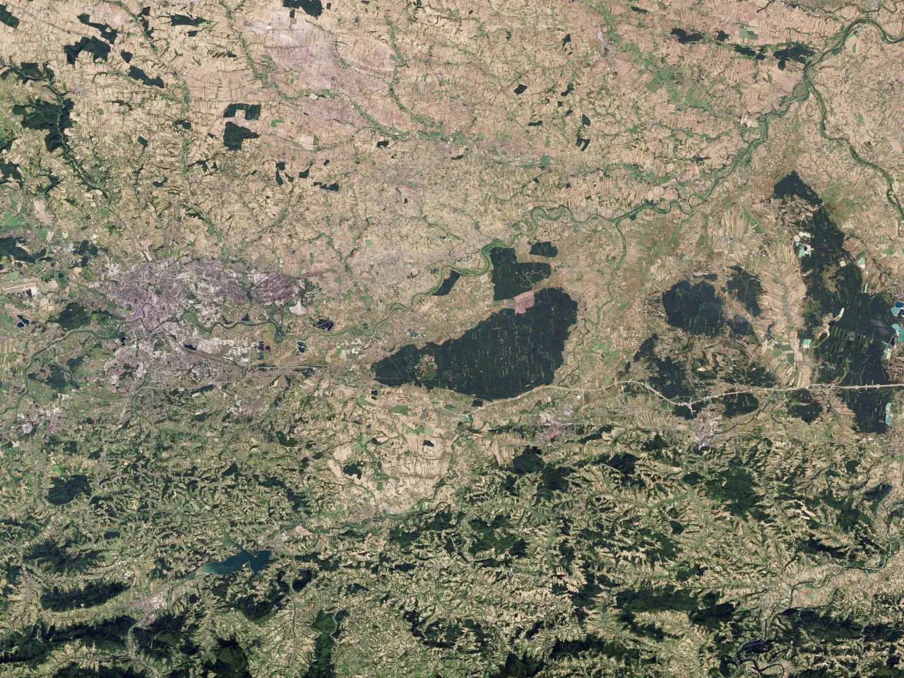Widok z lotu ptaka na krajobraz z polami uprawnymi i lasami, gdzie leżą Niepołomice. Widać wijącą się rzekę i zabudowania.