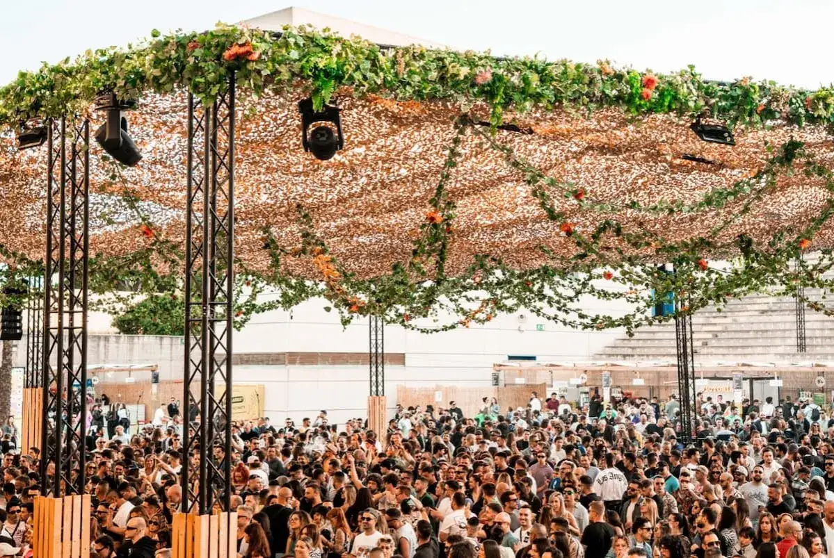 Multitud disfrutando de un evento al aire libre con una estructura decorada con vegetación y luces, similar a un **brunch electrónico en Málaga**.