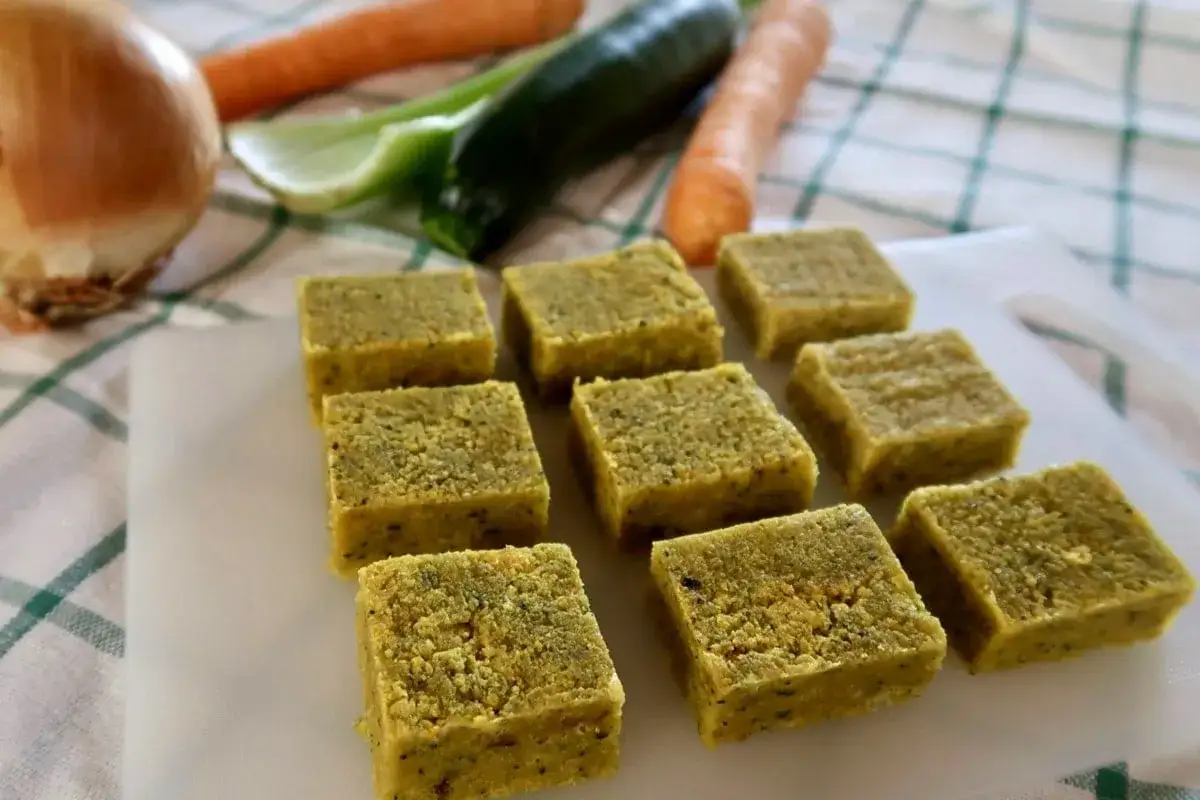 Cubetti di dado vegetale fatto in casa, pronti per insaporire le tue ricette. Sullo sfondo cipolla, carote, sedano e zucchina.